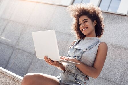 Young woman wearing overall shorts in the city street sitting browsing laptop relaxed joyfulの写真素材