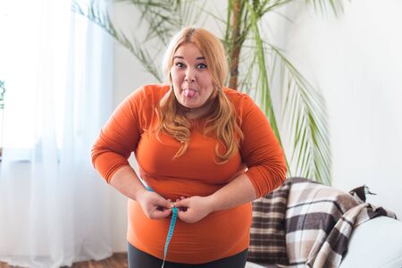 Chubby woman sport at home standing measuring waist showing tongue playfulの写真素材