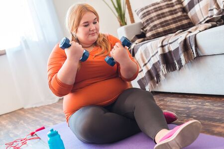 Chubby woman sport at home sitting on mat holding dumbbells thoughtfulの写真素材