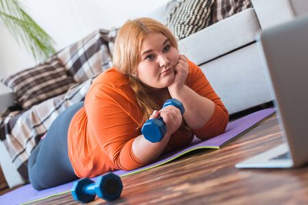 Chubby woman sport at home lying on mat watching video on laptop bored raising dumbbellの写真素材