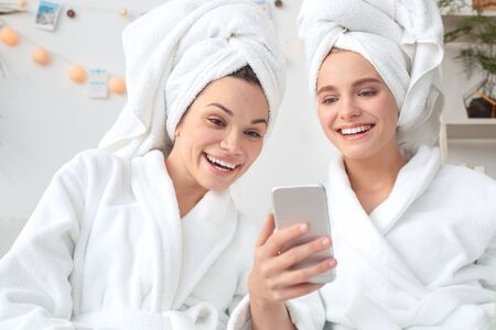 Friends together at home beauty care sitting in towels watching video on smartphone laughing happy close-upの写真素材