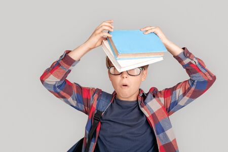 Freestyle. Young boy in glasses standing isolated on grey holding books on head surprised close-up