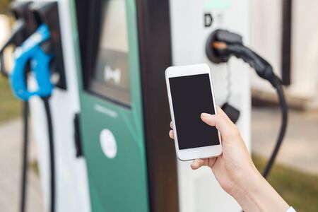 Cropped view of young adult woman hand holding smartphone with blank screen and copy space, paying for charging electric car, standing near city station with power cable supplyの写真素材