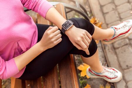 Young woman maintaining healthy lifestyle sitting on bench outdoors autumn season checking time on...