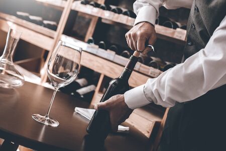 Senior man sommelier standing near cabinet opening wine bottle using corkscrew close-upの写真素材