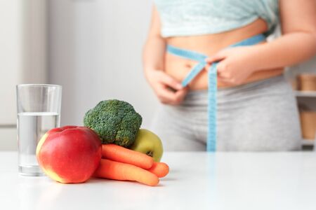 Young chubby woman on diet standing holding tape measure around waist near table with glass of pure water fresh vegetables and fruits in kitchen cclose-up blurred backgroundの写真素材
