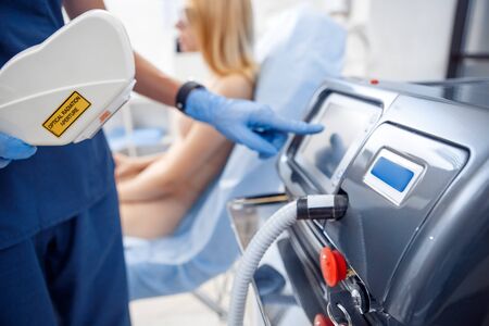 Young woman at beauty clinic cosmetology service sitting on medical chair blurred while doctor wearing gloves touching screen customizing photorejuvenation laser device close-upの写真素材