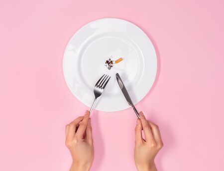 Person holding knife and fork eating cigarette butt from white plate isolated on pink background top view close-up killing habitの写真素材