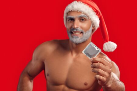 Christmas Freestyle. Young Santa Claus bare muscular upper body standing isolated on red showing condom to camera laughing playful blurredの写真素材