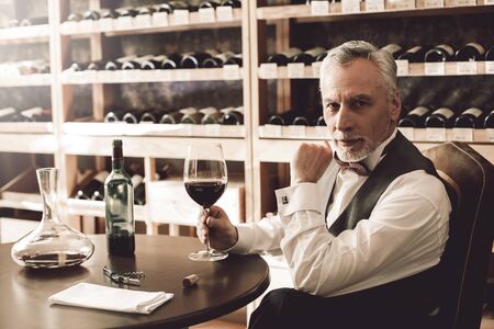 Sommelier Concept. Senior man sitting at table with glass of wine looking camera curiousの写真素材