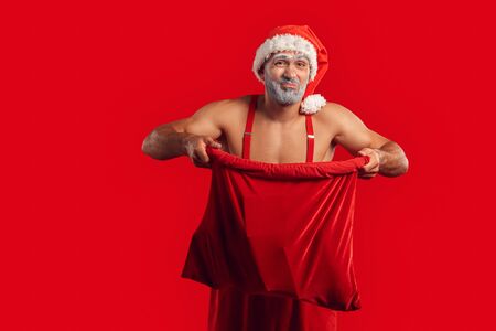 Christmas Freestyle. Young Santa Claus bare muscular upper body in hat standing isolated on red opening gift bag grimacing disgustedの写真素材