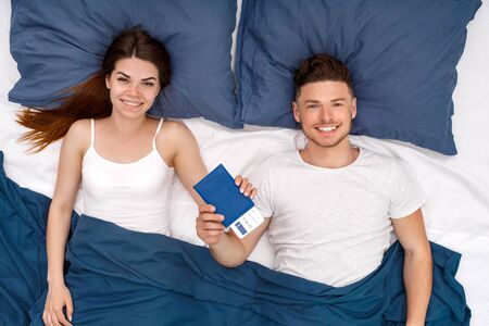 Bedtime. Young couple lying on bed under blanket top view with passport and ticket smiling cheerfulの写真素材