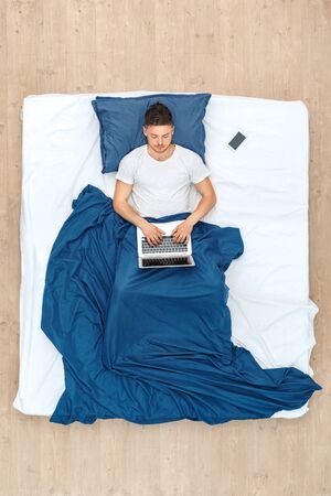 Bedtime. Guy lying on bed under blanket top view working on laptop concentratedの写真素材