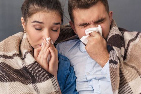 Feeling Sick. Young couple sitting on sofa covered with blanket studio isolated on grey blowing nose close-upの写真素材