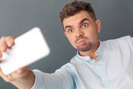 Freestyle. Young man studio standing isolated on grey taking selfie on smartphone grimacing to camera playful close-upの写真素材