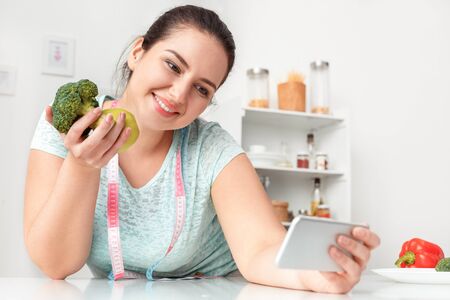 Body Care. Chubby girl leaning on table with tape measure around neck in kitchen taking selfie on smartphone posing to camera with broccoli and apple smiling cheerful close-upの写真素材