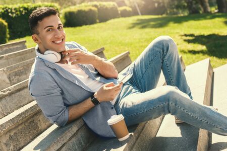 Outdoors Leisure. Stylish guy in headphones wearing watch sitting on stairs in park with cup of coffee smiling relaxedの写真素材