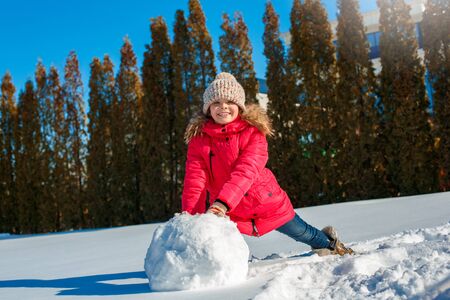 Winter vacation. Girl standing outdoors near house sitting rolling snowball for snowman smiling...