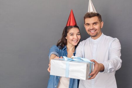 Celebration Concept. Young couple in birthday hat studio standing isolated on grey giving present to camera smiling friendlyの写真素材