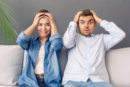 Relationnship Concept. Young couple sitting on sofa studio isolated on grey holding head shockedの写真素材