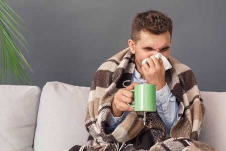 Feeling Sick. Young man sitting on sofa under blanket studio isolated on grey with cup of tea blowing his noseの写真素材