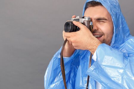 Photography Hobby. Young man in raincoat studio standing isolated on grey with film camera taking photos excited close-upの写真素材