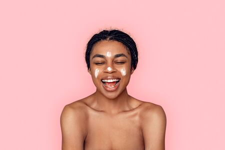 Beauty Concept. Young african woman isolated on pink having cream on face laughing cheerfulの写真素材