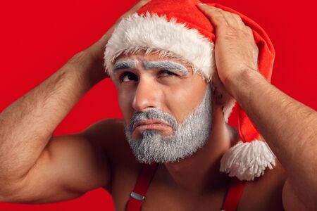 Christmas Freestyle. Young Santa Claus bare muscular upper body in hat standing isolated on red holding head upset close-upの写真素材