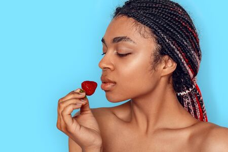 Beauty Concept. Young african woman isolated on blue looking at strawberry curious close-upの写真素材