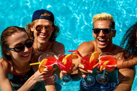 Summer Vacation. Young people in sunglasses swimming in the pool cheers with cocktails laughing...