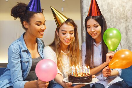 Young women having fun together indoors drinking wine celebrating birthday holding cake ready to blow the candles make a wish happyの写真素材
