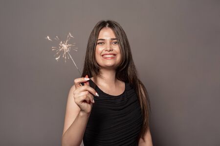 Freestyle. Young girl in dress standing studio isolated on grey holding sparklers smiling joyfulの写真素材
