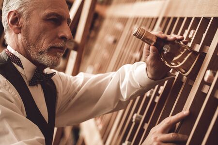 Sommelier Concept. Senior man standing taking bottle of white wine pensive close-up