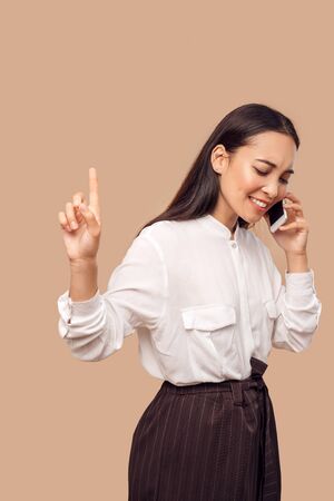 Freestyle. Young woman in shirt standing isolated on bage talking on smartphone showing silence gesture concentrated on callの写真素材
