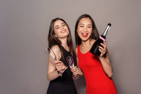 Freestyle. Young girls in dresses standing isolated on gray with champagne and glasses posing smiling excitedの写真素材