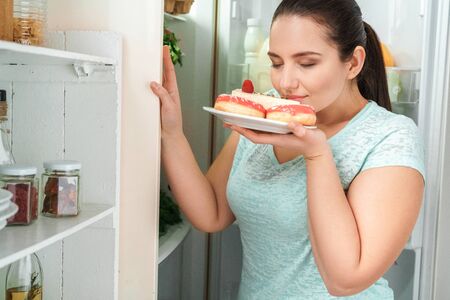 Body Care. Chubby girl standing in kitchen near fridge holding plate with doughnuts and cheesecake smelling joyful close-upの写真素材