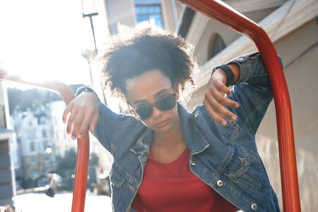 Outdoors Leisure. Young stylish african girl in denim jacket and sunglasses standing leaning on bar...