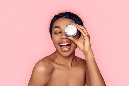 Beauty Concept. Young african woman isolated on pink covering eye with cream jar laughing cheerful