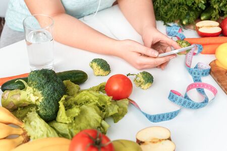 Young chubby woman wearing earphones standing in kitchen sitting at table with fresh healthy...
