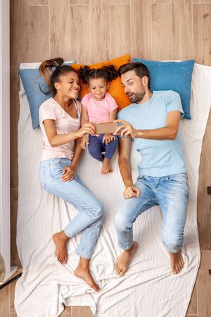 Mixed Race Family. Mother father and daughter lying on bed taking selfie on phone smiling cheerful top viewの写真素材