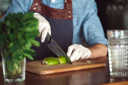 Professional bearded bartender wearing gloves standing at bar counter holding knife cutting lime on board for mojito close-upの写真素材
