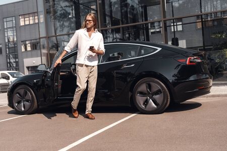Mode of Transport. Man with long hair going out of electric car with smartphone looking aside...