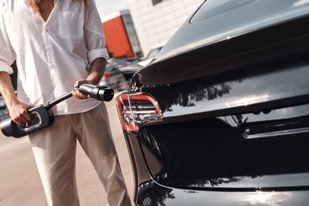 Young man traveling by electric car having stop at chraging station standing inserting charger close-upの写真素材
