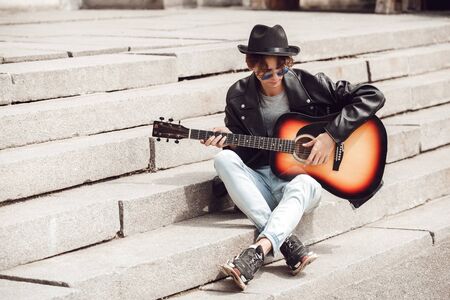Musician. Guy in hat and sunglasses sitting on stairs playing guitar pensive