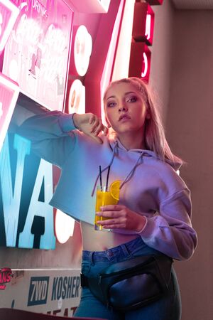 Nihgtlife. Teen girl leaning on wall isolated on club neon sign with cocktail posing to camera sassyの写真素材