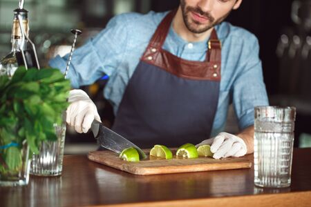 Drink Preparation. Bartender in gloves standing at counter cutting lime into cubes concentrated clsoe-upの写真素材