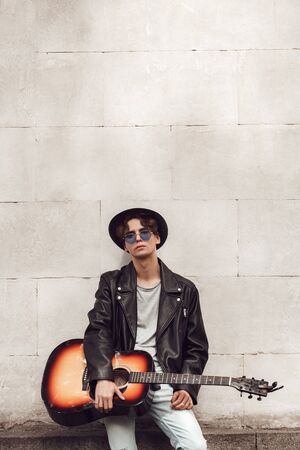 Musician. Guy in hat and sunglasses standing isolated on wall with guitar posing coolの写真素材