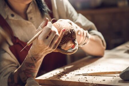 Craftsperson Concept. Young woman making pottery indoors sitting creating pattern on cup using modeling tool close-upの写真素材