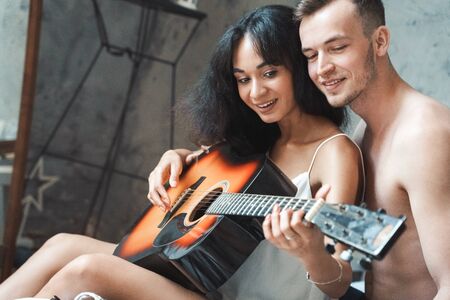 Mixed Race Couple. Young man teaching woman playing guitar sitting on bed together smiling joyful