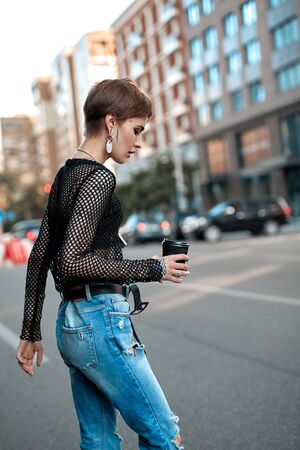 Urban Style. Young stylish woman walking outdoors on road with cup of coffee side viewの写真素材